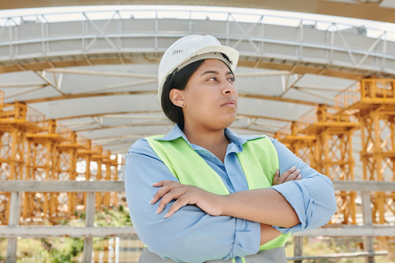 Young female engineer crossing her arms at a construction site, wearing a hardhat and reflective vest.